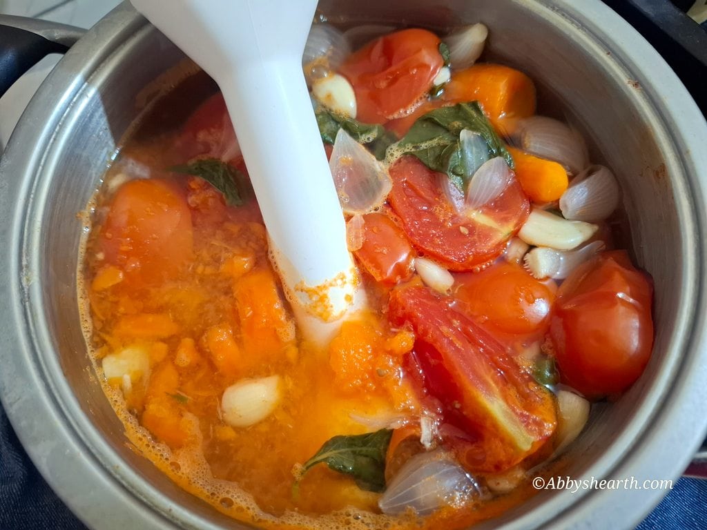 Homemade tomato carrot soup in a pot with a hand blender mashing it.