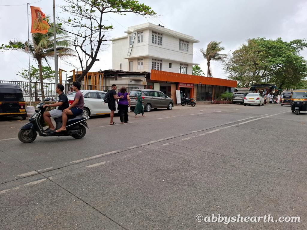 People and vehicles on a busy Mumbai street with a white cafe building in the background.