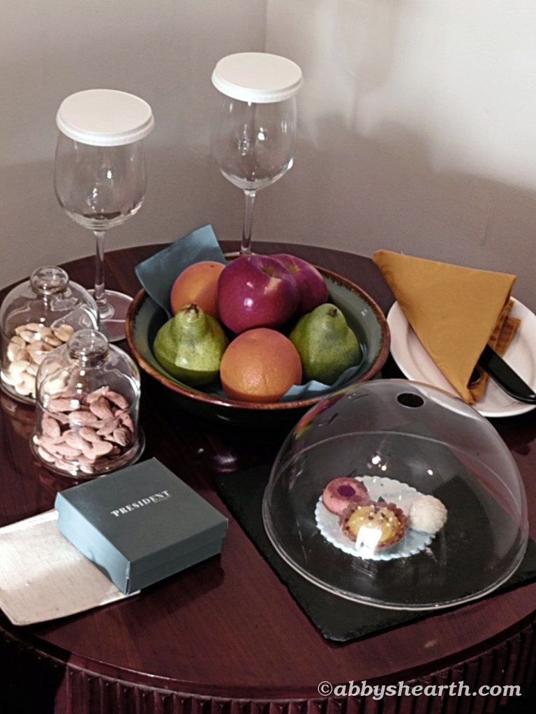Fruit bowl with apples, pears, and oranges on a dining table with glasses, nuts and complimentary desserts.