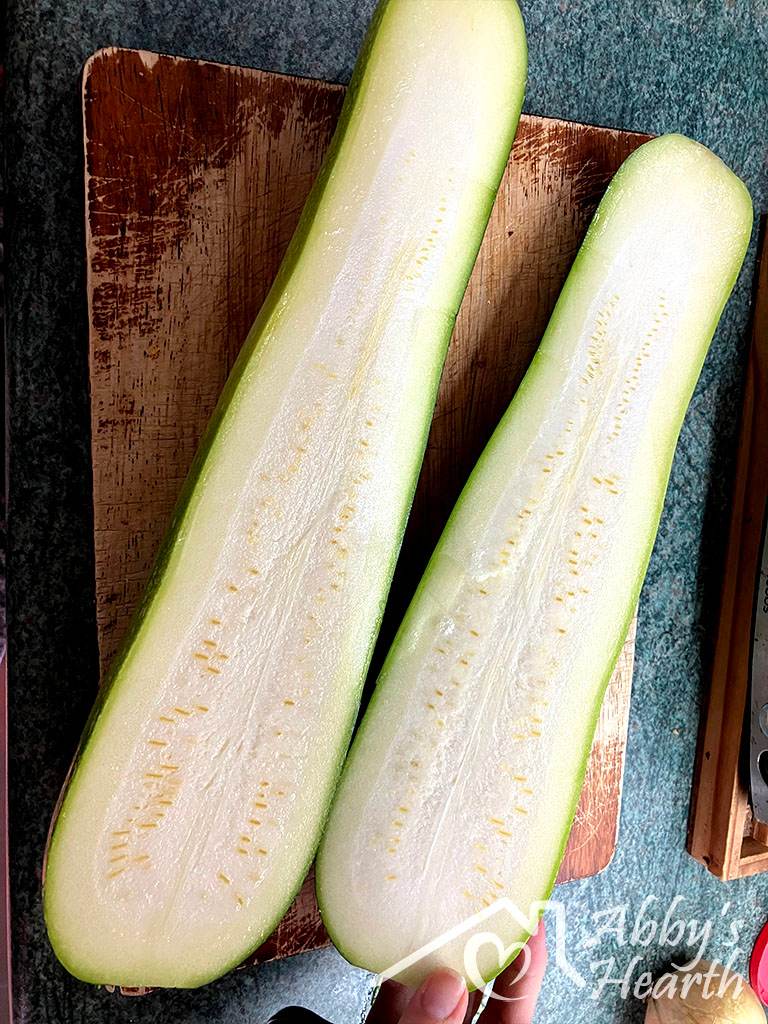 Two straight zucchini halves cut lengthwise on a wooden chopping board, seeds visible.