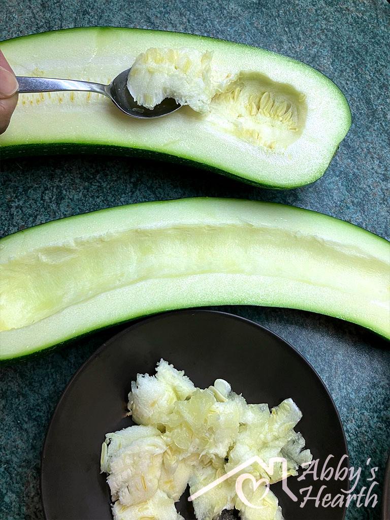 A spoon scooping pulp from a zucchini half, with the scooped pulp collected in a small dark bowl below.