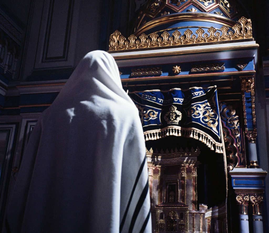 Person praying at a synagogue with a tallit and ornate ark in the background.