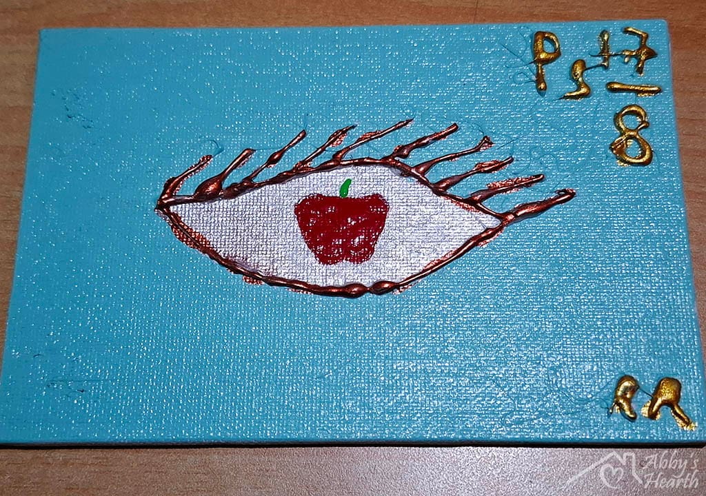 Glue art of an eye with an apple psalm 17 verse 8 on blue canvas.