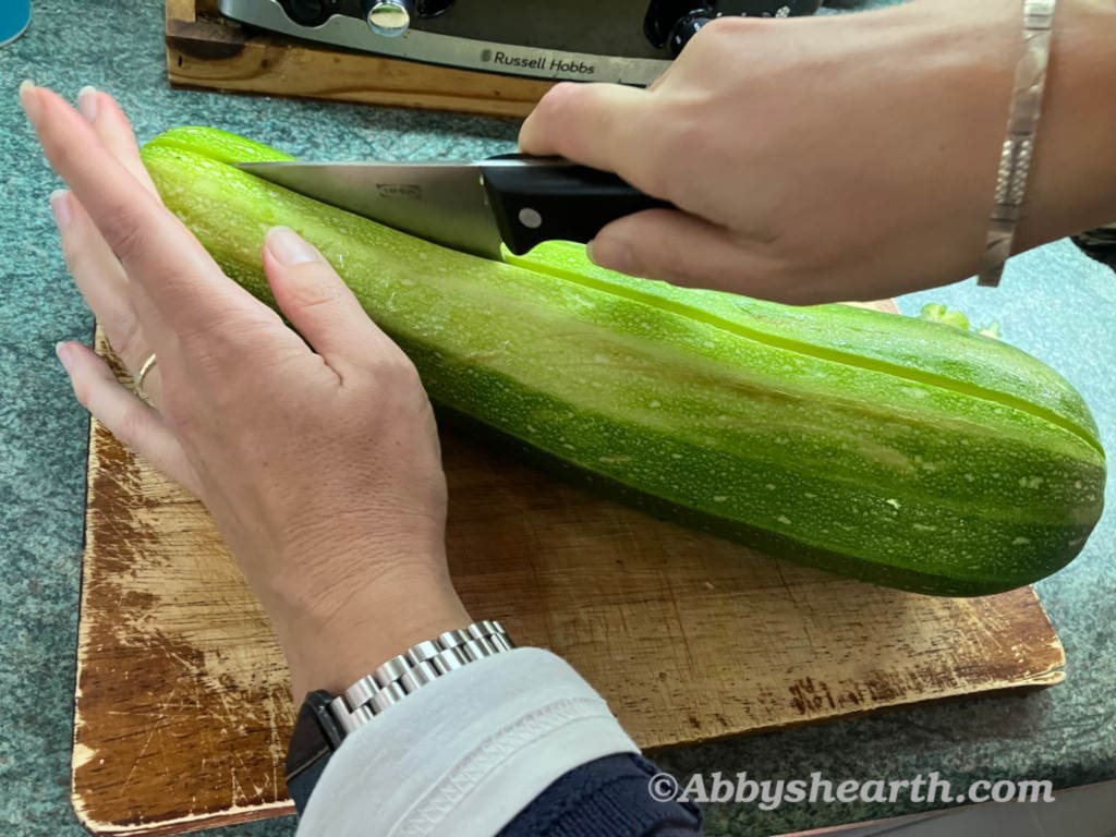 Hands cutting a zucchini lengthwise on a wooden chopping board.