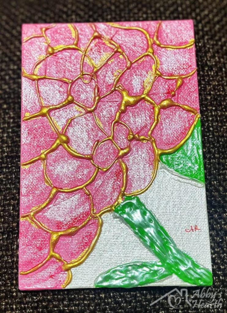 Pink and golden glue art flower with green stem and leaves.