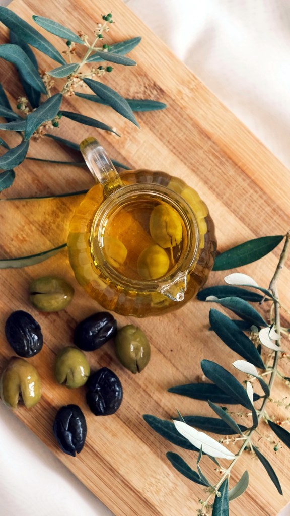 Jar of olive oil surrounded by olives and olive leaves.