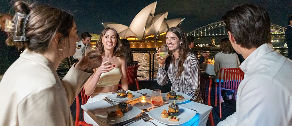 Smiling people on a Sydney dinner cruise with Opera House & Harbour Bridge in the background.