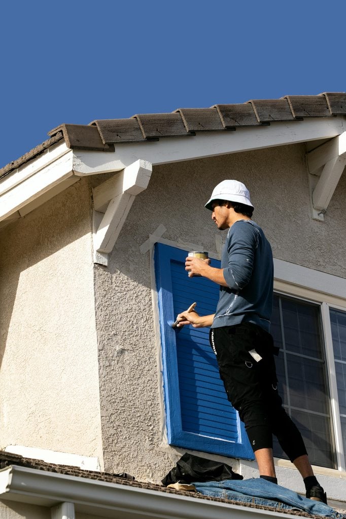 Man standing on a roof painting a window.