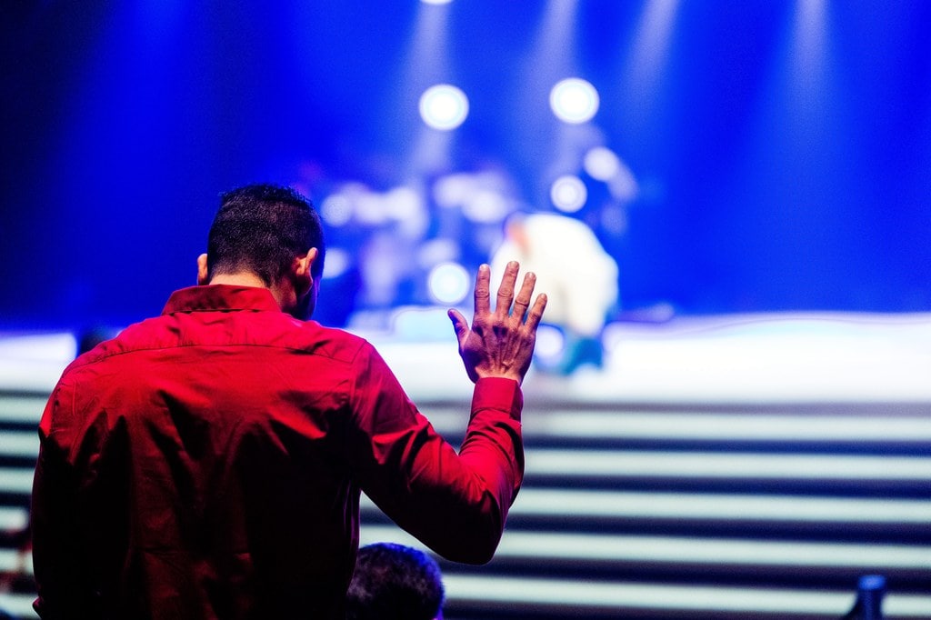 Man in red shirt in front of a stage praying with hands raised.