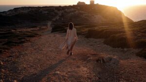 Woman in long dress walking towards the sunset on a cliff.