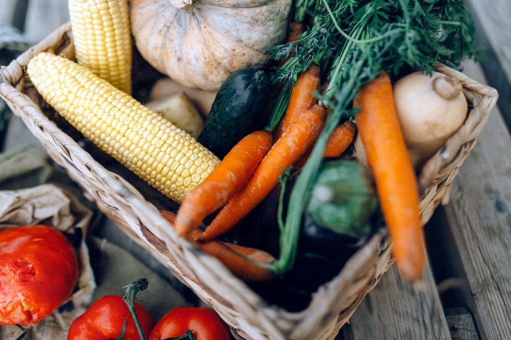 Assorted fresh vegetables including carrots, corn, zucchini, and tomatoes in a basket.