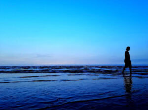 Person walking alone on the beach during sunset with calm waves and blue sky.