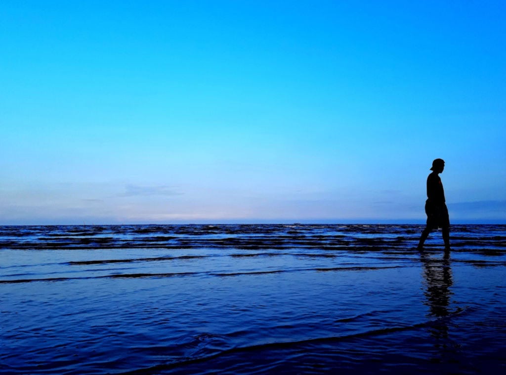 Person walking alone on the beach during sunset with calm waves and blue sky.