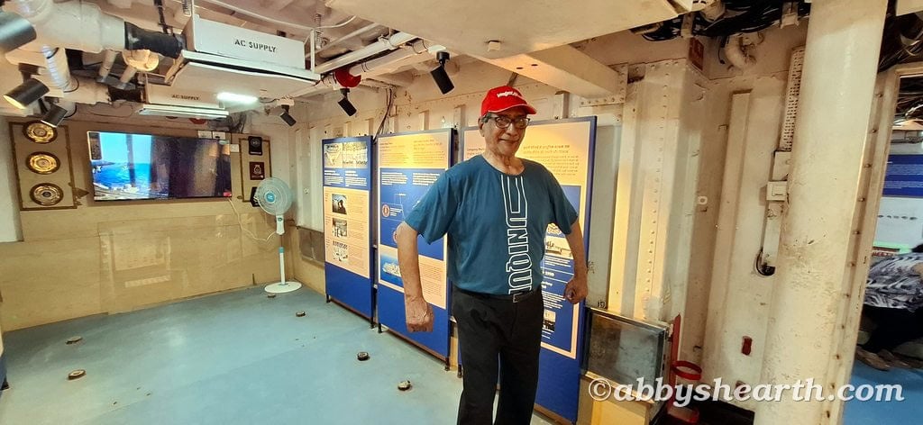 Senior man at Diu INS Khukri showing muscles with informational displays in background.