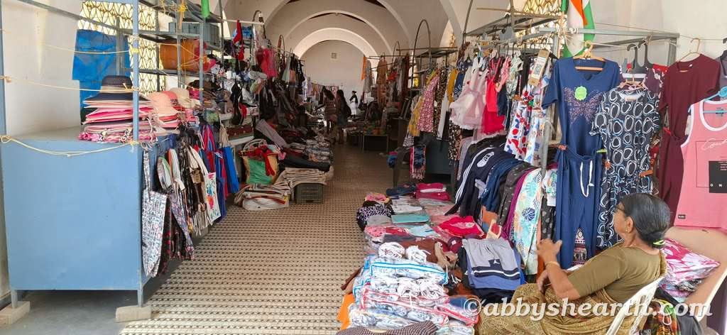 Indoor market with clothing and fabric stalls, shoppers browsing, vibrant display.