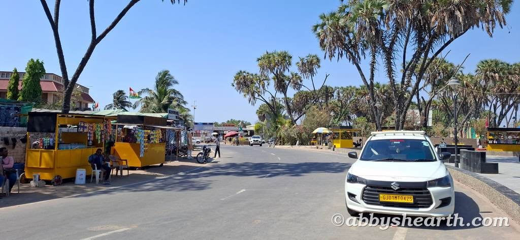 Street view in Diu with cars, vendors, and hoka palm trees under clear sky.