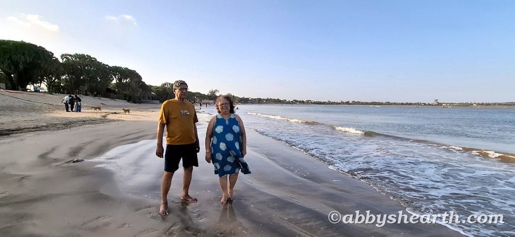 Senior parents walking along Nagoa beach in Diu with ocean waves and clear sky in background.