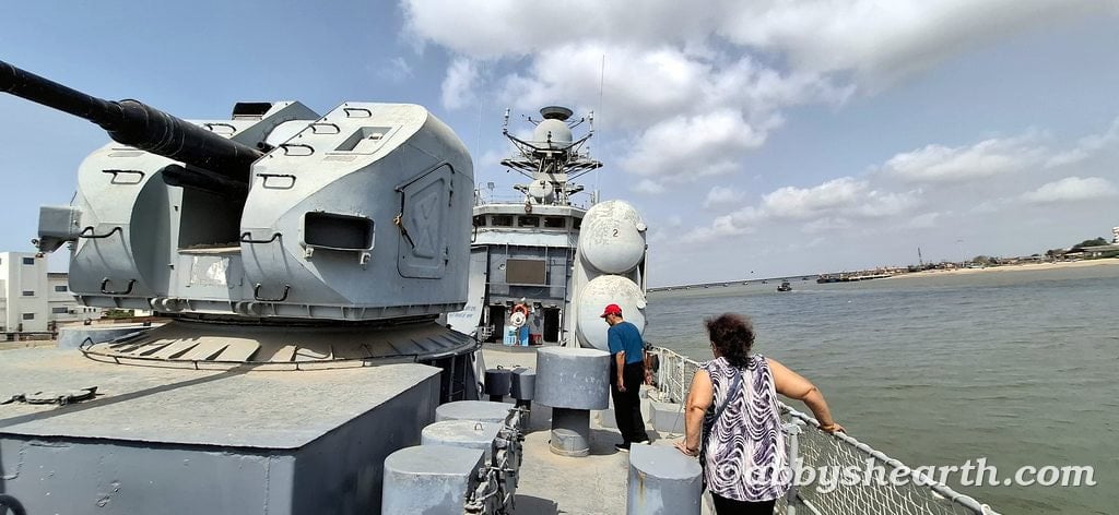Tourists exploring a naval ship in Diu harbor with a large gun turret.