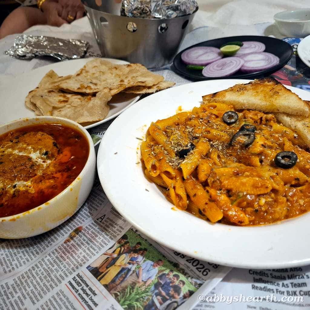 Plate of pasta with black olives, naan bread, and spicy curry on newspaper.
