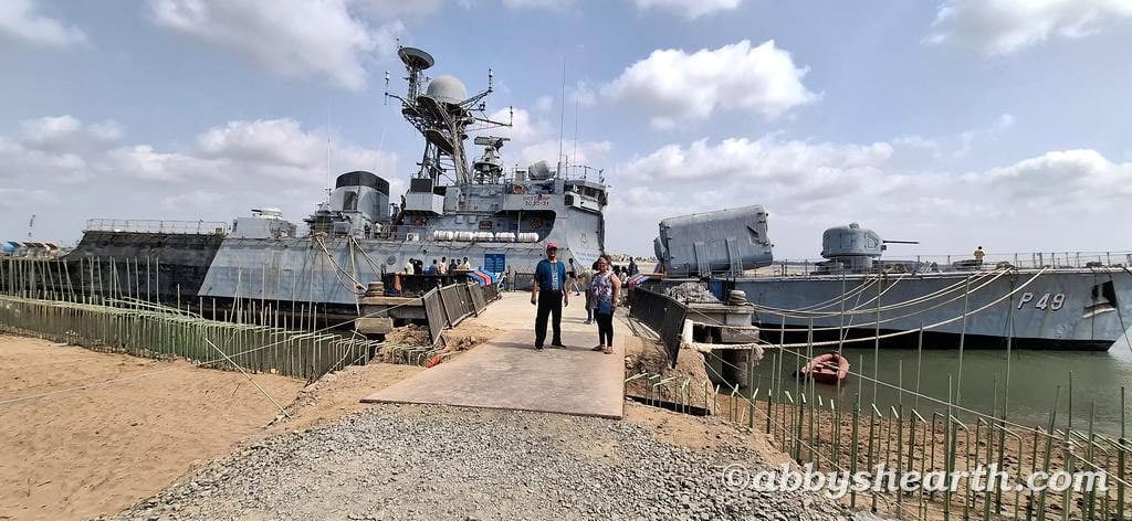 Visitors walking on a pier towards a historic warship at Diu port with blue sky and clouds.