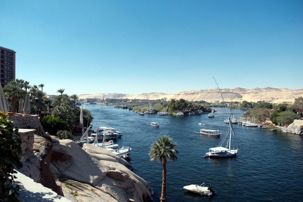Scenic view of the Nile River with boats, palm trees, and desert hills in the background.
